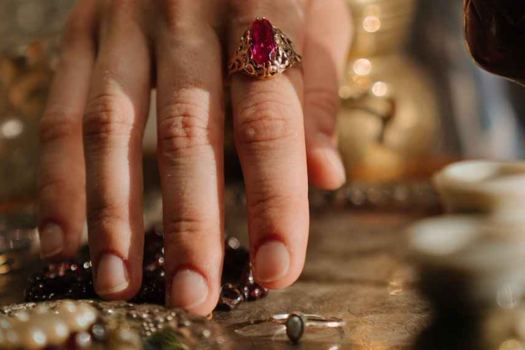 A close-up photo of a woman's hand wearing a red gemstone ring and holding other pieces of gemstone jewelry