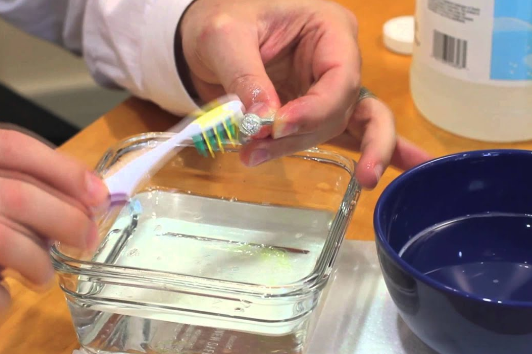 The image shows a professional hand engaged in the process of cleaning. An image of a diamond ring being gently brushed with a soft brush