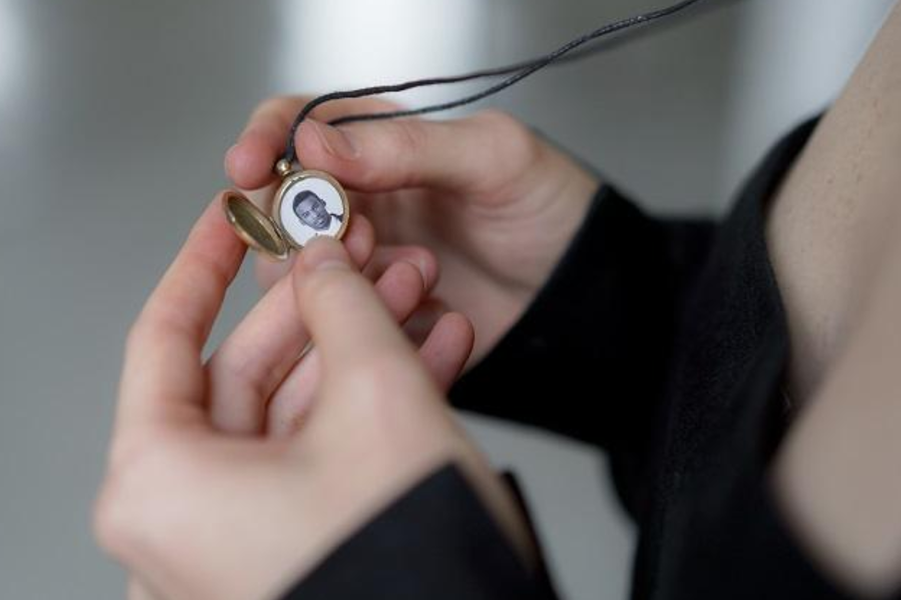 A photograph of a woman's hand holding a necklace with a birthstone pendant. The pendant features a picture of a man