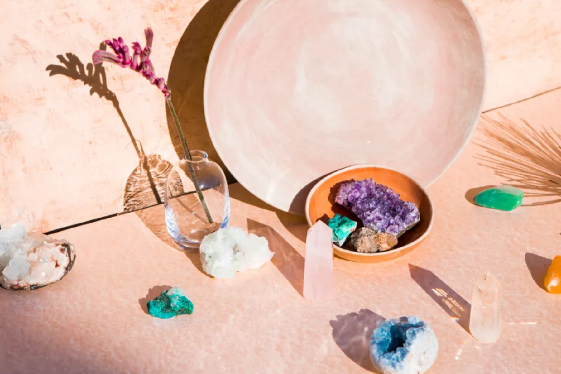 A pink scene with various crystals in an orange bowl, other stones outside, and a flower in a glass jar