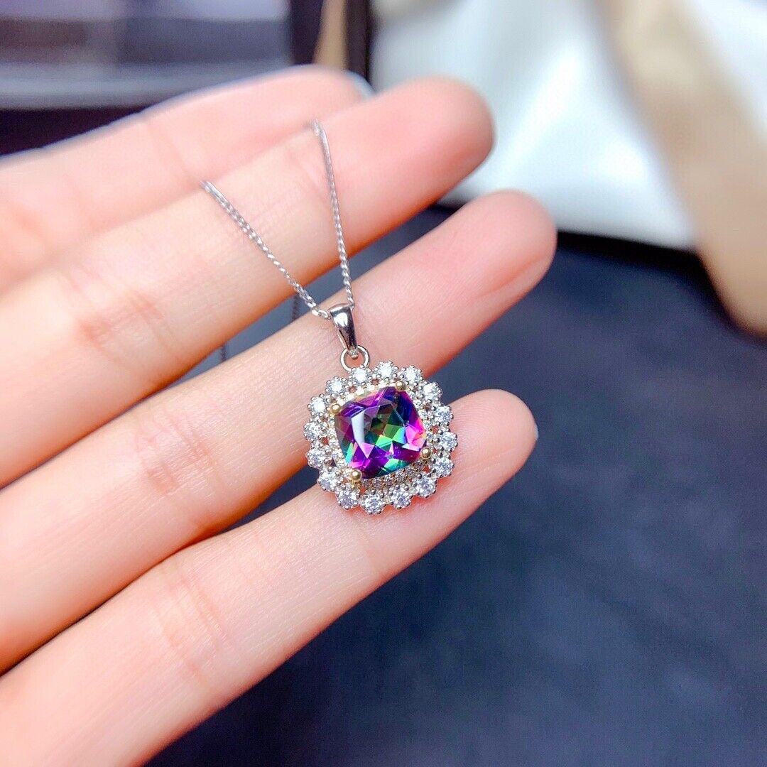 A hand holds a square-cut mystic topaz pendant surrounded by a halo of small gemstones, hanging from a thin silver chain.